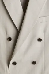 Blazer de doble botonadura en color beige, con solapas de pico, bolsillos delanteros con solapa y botones oscuros en contraste. Presenta un corte estándar con abertura doble en la parte trasera y forro interior.