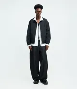 Campera masculina de corte boxy en color beige, con cuello inglés, cierre frontal de cremallera, mangas largas con puños elásticos y ruedo con elástico. Presenta bolsillos laterales y detalles de recortes en el diseño.