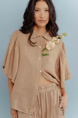 Camisa de lino color tostado, con cuello camisero y cierre frontal de botones. Se caracteriza por sus mangas cortas amplias y voluminosas que aportan un diseño fresco y liviano.