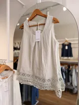 Blusa sin mangas de color beige, confeccionada en mezcla de lyocell y lino, con diseño de pliegues en el frente y detalle de encaje tipo puntilla en el ruedo.