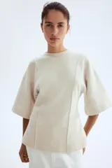 Top de punto fino con cuello redondo, hombros caídos y mangas al codo. Presenta costuras decorativas verticales en el frente y un bajo levemente acampanado con corte holgado.