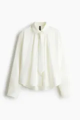 Camisa de manga larga con cuello clásico y botonadura frontal. Presenta una corbata extraíble a juego, puños con cierre de botón y un sutil detalle bordado en el pecho. El diseño incluye un canesú con pliegue en la espalda y un bajo redondeado con aberturas laterales.