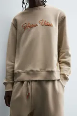 Sudadera de algodón color beige claro, corte relajado, cuello redondo y manga larga. Presenta bordado en relieve en el frente con texto en cursiva de color cobre.