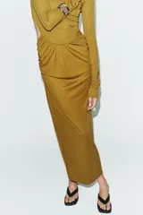 Vestido largo color mostaza con cuello alto, manga larga drapeada y detalles cut out laterales. Presenta frunces en la cintura y la espalda, y bajo recto.