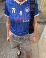 Camiseta de fútbol de manga corta en color azul con diseño estampado abstracto y cuello en V rosado con detalles blancos a juego con los puños. Presenta inscripciones y escudos deportivos en el pecho.