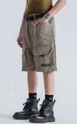 Bermuda técnica color arcilla (marrón claro/beige) de calce oversize, confeccionada en poliéster liviano. Presenta múltiples bolsillos cargo con tapa y un cinturón de ajuste con hebilla plástica negra. Incluye detalles reflectivos.