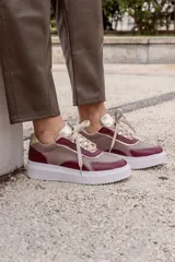 Championes urbanos con diseño de paneles en cuero color beige, bordó y detalles metalizados dorados en la lengüeta y talón. Presentan costuras decorativas visibles en contraste y suela de plataforma blanca con textura acanalada.