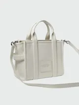 Cartera tipo tote bag Marc Jacobs modelo The Leather Small Tote Bag, color negro, confeccionada en cuero graneado con el texto 'THE TOTE BAG' grabado en relieve al frente. Cuenta con asas superiores, correa ajustable y desmontable, y herrajes metálicos dorados.