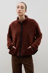 Campera de sherpa color marrón con cierre frontal, cuello alto y bordado de la marca en el pecho. Presenta detalles de ribetes en color negro en los bordes y bolsillos laterales.