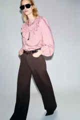 Camisa de manga larga con cuello subido, cierre frontal con botones y detalle de volantes en el pecho. Presenta un diseño a rayas verticales y puños elásticos.