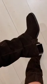 Bota de caña alta de gamuza en color marrón chocolate, con taco cuadrado y puntera cuadrada. Cuenta con un diseño holgado en la caña y detalle de trabilla decorativa en el tobillo.