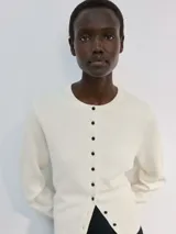 Cardigan de punto color blanco marfil, con cuello redondo, manga larga y cierre frontal con pequeños botones negros en contraste.