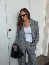 Conjunto de dos piezas compuesto por un blazer y un pantalón de corte sastrero, confeccionados en tela de algodón con estampado de rayas verticales blancas sobre fondo gris. El blazer presenta solapas clásicas y cierre frontal, mientras que el pantalón es de tiro medio y pierna recta.