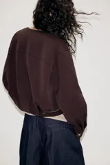 Chaqueta estilo bomber de color marrón oscuro, con cuello redondo, manga larga y cierre frontal con cremallera. Presenta bolsillos delanteros de vivo y un diseño con bajo fruncido.