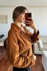 Campera tipo bomber de corte corto, confeccionada en gamuza color camel. Presenta cuello solapa, cierre frontal con botones, forro interior y detalles decorativos de pasamanería trenzada a lo largo de las mangas.