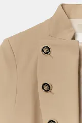 Blazer entallado de sarga color beige con cuello subido y escote en pico. Presenta manga larga con hombreras, detalle de botones metálicos decorativos en el frente y cierre frontal con gancho metálico. Incluye falsos bolsillos de vivo y abertura en la espalda.