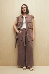 Chaleco color beige con cuello camisero, sin mangas, cierre frontal con botones y cinturón de lazo. Presenta bolsillos de parche en el pecho y pinzas verticales en el frente.