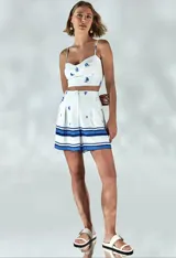 Short sastre blanco de tiro alto, corte recto y diseño con pliegues, con estampado de pequeños barcos azules y franjas horizontales en tonos azules en el bajo. Cuenta con bolsillos y cierre frontal con botón cubierto.