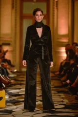 Blazer tipo smoking de terciopelo negro con acabado brillante y detalles estampados en dorado. Presenta solapas clásicas, cierre frontal cruzado mediante un lazo y mangas largas con cierres incorporados. El diseño es entallado y sofisticado.