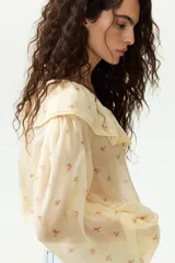 Blusa de gasa arrugada de corte holgado, en tono amarillo claro con estampado floral. Cuenta con escote en V profundo, un amplio volante ribeteado con encaje alrededor del cuello y cordones finos para atar al frente. Mangas largas con puños elásticos.