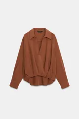 Camisa de lino color beige, con cuello solapa, escote en V y mangas largas. Presenta un diseño de bajo asimétrico con pliegues delanteros que generan un efecto abullonado.