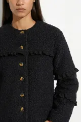 Campera de tela bouclé color negro, con diseño de cuello redondo y cierre frontal mediante botones. Presenta detalles de volados en tela agamuzada en el pecho y en las mangas.