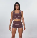 Conjunto deportivo de dos piezas color marrón, compuesto por un top deportivo con cuello redondo y costuras decorativas en contraste, y un short de tiro alto con costuras laterales a juego. El top presenta el logo 'bk.' estampado en el frente.