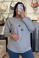 Buzo tejido de punto en color beige, con diseño de flores aplicadas en tono gris oscuro y detalles de brillos plateados que irradian desde las flores. Presenta cuello redondo amplio, mangas largas con caída holgada y corte recto.