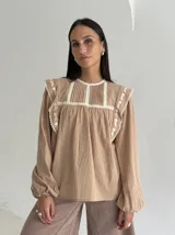 Blusa color beige de algodón con cuello redondo, mangas largas con puños elásticos y detalles de puntilla y bordados en el frente y hombros.