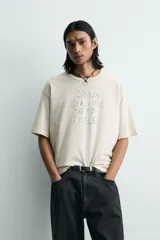 Camiseta de corte holgado y estilo cropped, color beige con efecto lavado. Presenta cuello redondo, mangas cortas y una estampa frontal con texto en azul que simula trazos de rotulador.