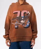 Buzo con capucha color marrón con efecto teñido/desgastado, estampado frontal grande con el número "76" con diseño de bandera estadounidense, un águila calva y una cinta con el texto "AMERICAN LOVE". Tiene bolsillo canguro y tres pines decorativos prendidos en la zona del hombro.