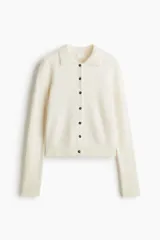 Cárdigan corto de punto suave color crema, con cuello camisero, abotonadura frontal con botones marrones y remate acanalado en puños y bajo.