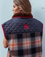 Chaleco sin mangas de diseño híbrido que combina paneles de tela acolchada capitoné en color azul marino con secciones de tejido de lana a cuadros en tonos naranja, gris y negro. Presenta cuello con forro de piel sintética marrón, cierre frontal con cremallera y ribetes en color rojo vibrante. Incluye bolsillos delanteros amplios con cierre.