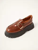 Mocasines de cuero color marrón con plataforma dentada negra. Presentan una tira frontal con hebilla metálica dorada y costuras decorativas en contraste.