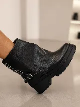 Bota corta de cuero negro con diseño de polaina superpuesta, detalle de tachas metálicas en el lateral y correa con hebilla ajustable. Cuenta con suela track de goma y taco cuadrado.