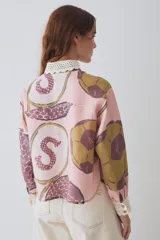 Camisa de lino blanca con estampado de motivos deportivos, cuello y puños tejidos a mano en crochet azul con borde rojo. Presenta cierre frontal con botones y un bolsillo en el pecho.
