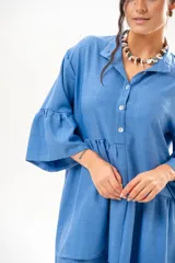 Vestido corto azul, estilo camisero, con cuello camisero, cierre frontal con botones y mangas acampanadas con volados. Presenta un diseño holgado con cortes fruncidos en el cuerpo.