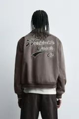 Cazadora estilo varsity de tejido efecto ante en color beige. Presenta cuello solapa, cierre frontal con botones a presión y puños y ruedo con acabado en rib a rayas negras. Incluye parches bordados decorativos en el pecho y la manga, además de bolsillos laterales de doble vivo.
