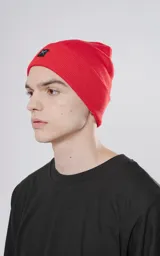 Gorro de tejido tipo beanie, color rojo, con borde doblado y un pequeño parche negro bordado con un símbolo de tijeras cruzadas en blanco.