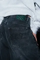 Pantalón de jean estilo baggy color negro con efecto lavado, corte holgado y diseño de cinco bolsillos. Presenta un pequeño dije decorativo en la cintura y detalle de logo metálico en el frente.