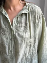 Camisa verde claro de corte oversize, con mangas tres cuartos abullonadas, escote en V con cuello camisero y cierre frontal parcial de botones. Cuenta con delicados bordados florales tono sobre tono distribuidos por toda la prenda.