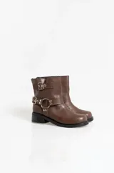 Botas de caña baja en cuero sintético color marrón, con diseño de estilo biker que incluye dos correas horizontales con hebillas metálicas y una argolla decorativa en el lateral. Posee suela y taco bajo de color negro.