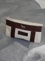 Sobre tipo cartuchera de lona naranja con detalles y apliques en cuero marrón. Presenta un aplique metálico en forma de caballo y un parche con el logo de la marca grabado en el frente.