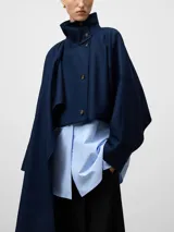Cazadora corta de gabardina en color azul marino, con cuello chimenea y cierre frontal de botones. Presenta un diseño estructurado con mangas amplias y una pieza de tela integrada que cae lateralmente a modo de capa o chal.