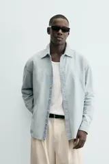 Camisa de denim de algodón, color celeste claro lavado, de corte relajado (relaxed fit) con cuello solapa, manga larga terminada en puño con botón y cierre frontal de botonadura. Presenta un pequeño bordado a contraste en el pecho.