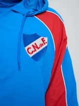 Campera deportiva Umbro del Club Nacional de Football, modelo Venti, para hombre. Diseño en color blanco con mangas azules y detalles de líneas rojas. Cuenta con capucha con cordón ajustable, cierre frontal y bolsillos. Presenta el logo de Umbro y el escudo del club bordados en el pecho.