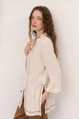 Chaqueta beige de lino con mangas 3/4, cuello mao y detalles de costuras decorativas oscuras en contraste.