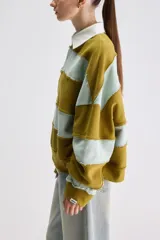 Buzo de estilo rugby con diseño oversize, confeccionado en bloques de color verde militar y celeste con costuras expuestas y deshilachadas. Cuenta con cuello camisero blanco de popelina y cartera de botones frontal oculta, además de mangas largas y puños acanalados.