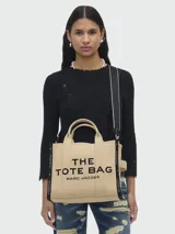 Bolso tote Marc Jacobs modelo The Jacquard, confeccionado en tejido de lona negro con detalles en blanco. Presenta el logo de la marca y la inscripción 'THE TOTE BAG' en el frente, con doble asa superior y correa ajustable extraíble.