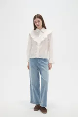 Camisa de algodón blanca con cuello camisero, cierre frontal de botones y volados bordados en el frente y hombros. Presenta mangas largas con puños acampanados y detalles bordados en el ruedo.